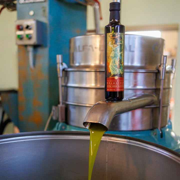 Bottle of olive oil being poured into a large metal container with industrial equipment in the background.